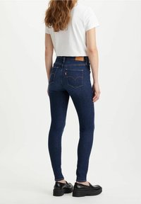 Jeans skinny blu scuro a vita alta in denim, con due tasche posteriori e un'etichetta in pelle color cammello sulla vita.