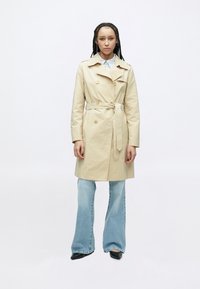 Béžový trenčkot s dvojradovým zapínaním na gombíky, opaskom v páse a klasickým golierom. Kombinovaný so svetlomodrými zvonovými džínsami a čiernou obuvou.