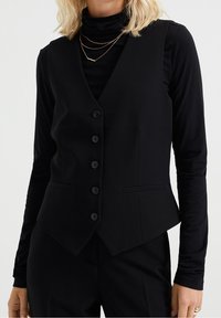 Svart skräddarsydd väst med v-ringning, sex knappar och två sidfickor. Slätt tyg med en åtsittande form och längre bakstycke.