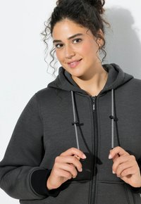 Szary hoodie na zamek z kontrastowymi czarnymi wykończeniami i sznurkami. Ma zamek z przodu, kieszenie oraz miękki, teksturowany materiał.