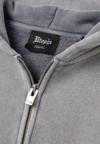 Grijze ritssluiting hoodie gemaakt van zachte stof met een fleece voering. Voorzien van een zilveren ritssluiting en een zwart etiket met de tekst "Utopia" en maat XS.