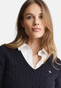 Marineblauer grob gestrickter Pullover mit V-Ausschnitt aus weichem Material, über ein weißes Hemd mit Kragen getragen, mit dezent gesticktem Logo.