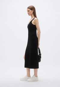 Čierne midi šaty bez rukávov s výstrihom do V, s hladkou textúrou a rozparkom na boku, doplnené bielymi teniskami a čiernou taškou.