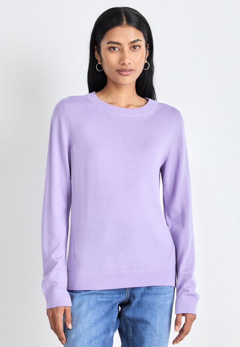 Suéter de malha de manga longa numa tonalidade lilás clara, com decote redondo, punhos e barra canelados; ajustado ao corpo com detalhes de fenda lateral. Combinado com jeans azuis.