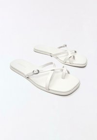 Sandales plates blanches avec des bouts carrés, des fines brides croisées et une petite boucle argentée sur une surface blanche propre.