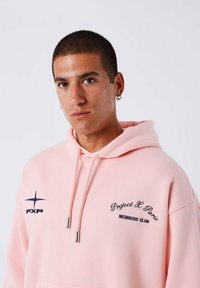 Jonge man met kort haar en oorringen, gekleed in een lichtroze hoodie met de tekst "Project X Paris Members' Club" op de borst.