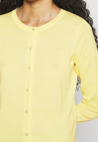 Ljusgul cardigan i mjukt material, med rund halsringning och fem pärlemorknappar fram för stängning.