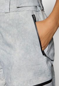 Gaiši pelēki īsie šorti ar režģa rakstu un gludu tekstūru. Iespējams, ar rāvējslēdzēja kabatu un melnām akcentām gar šuvēm.