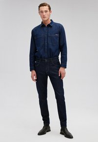 Denim shirt in dark blue with button-down collar, chest pockets, and long sleeves. Paired with dark blue denim jeans and black ankle boots.