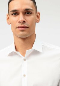 Close-up of a man with short dark hair wearing a white collared button-up shirt, looking straight ahead with a neutral expression.