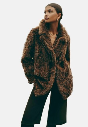 Pruun faux-furi jope laia kraega, suuruse ülekasvuga, tekstuurse pinna ja nööbikinnitustega, kantud tumeda särgi ja pükste kohal.