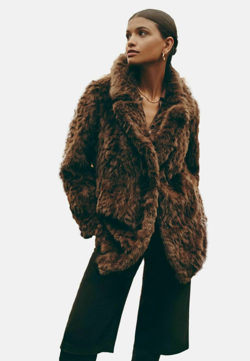 Brūns mākslīgās kažokādas mēteli ar plašu apkakli, pārliecinošu piegriezumu, teksturētu virsmu un pogu aizdari, vilkts uz tumša krekla un biksēm.