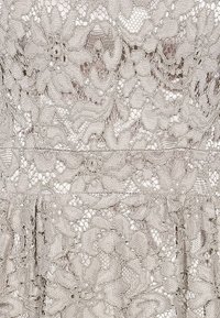 Tissu en dentelle beige avec des motifs floraux et géométriques complexes, comportant des bords festonnés et une texture transparente et délicate.
