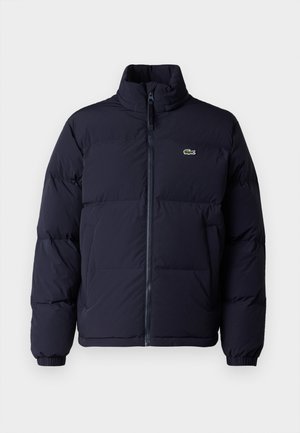 Casaco puffer azul-marinho com um colarinho alto, fecho de zíper e punhos elásticos. Apresenta um pequeno logótipo verde no lado superior esquerdo do peito.