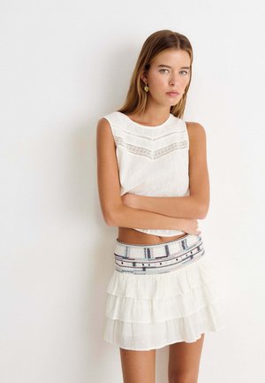 Jeune femme aux longs cheveux portant un haut blanc sans manches et une jupe à volants avec une ceinture brodée, debout devant un mur blanc uni.