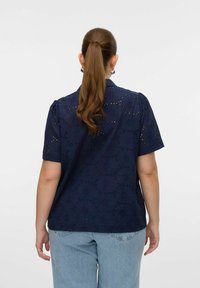 Camicia blu navy con maniche corte a palloncino, caratterizzata da motivi floreali tagliati al laser, realizzata in tessuto traspirante, indossata con jeans azzurri.