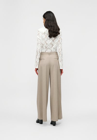 Γυναίκα με μακριά σκούρα μαλλιά, που φοράει λευκό πουκάμισο μακρυμάνικο δαντέλα με γιακά και μπεζ φαρδιά παντελόνια, από πίσω.