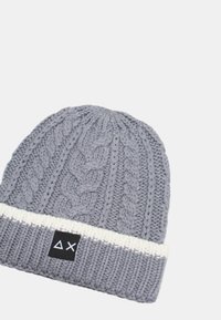 Berretto grigio lavorato a maglia con motivo a treccia, polsino a coste color crema e un'etichetta nera con simboli geometrici bianchi.