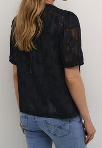 Blouse noire transparente à texture florale, manches courtes bouffantes et coupe décontractée. Associée à un jean bleu clair pour un look décontracté.