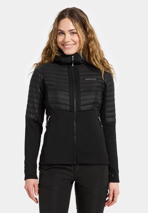Femme souriante avec de longs cheveux ondulés portant une veste noire zippée avec des épaules rembourrées et des poches latérales zippées, se tenant contre un fond blanc.