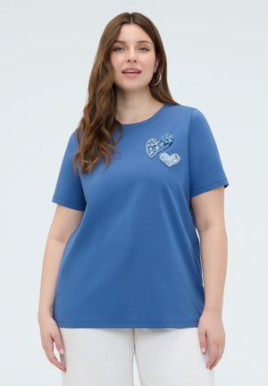 Kvinde iført en blå kortærmet skjorte med to udsmykkede hjertepatches på brystet og hvide bukser, stående mod en ensfarvet baggrund.