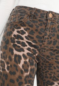 Jeans de denim marrón con un patrón de estampado de leopardo negro, con una textura suave, cinturilla y detalles de botones en color cobre.