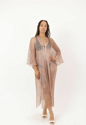 Vrouw met lange vlechten staat in een beige opengebreide, met franjes afgewerkte cover-up over een zwarte outfit en witte hak-sandalen tegen een effen achtergrond.