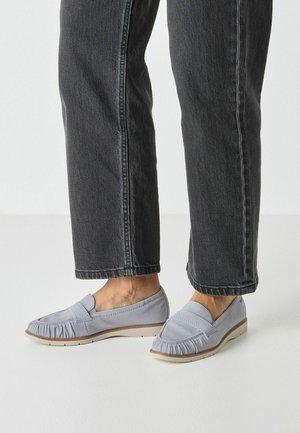 Mocassins gris avec une texture froncée, semelle plate et semelle intérieure rembourrée, associés à un jean sombre. Design minimaliste et matière douce.