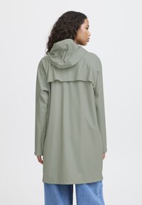 Impermeabile lungo di colore verde chiaro con cappuccio, dotato di vita cinchata, chiusure a pressione e un design a strati sulla schiena. Il tessuto sembra liscio e impermeabile.