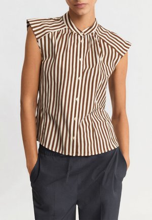 Blusa senza maniche a righe verticali marroni e bianche con bottoni frontali, indossata con pantaloni blu scuro con le mani nelle tasche.