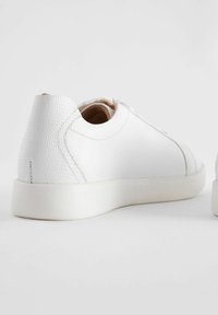 White leather sneakers with textured heel, round toe, flat rubber sole, and subtle stitching details. Minimalist design with clean lines.