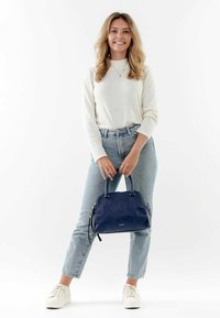 Navyblaue Lederhandtasche mit strukturiertem Design, doppelten Griffen und einem Reißverschluss. Das Modell trägt helle Jeans und einen cremefarbenen Pullover.