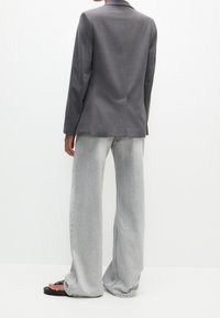 Blazer gris sur mesure avec une texture lisse et des revers crantés, associé à un jean gris clair à jambes larges et des tongs noires.