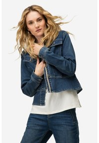 Denim takki keskisinisessä, lyhyessä mallissa nappikiinnitys, rintataskut ja vaaleat tikkaus; käytetty valkoisen paidan päällä.