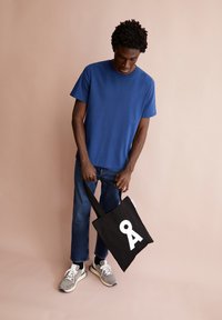 T-shirt en coton bleu, jean droit en denim, baskets grises, tenant un sac fourre-tout noir avec un design graphique blanc. Fond neutre.