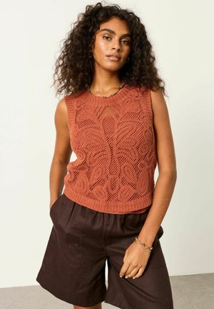 Frau mit lockigem Haar, die ein ärmelloses, rostorangefarbenes Stricktop und dunkelbraune Shorts mit hoher Taille trägt, die Hände in den Taschen.