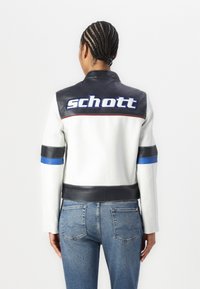 JAQUETA DE MOTOQUEIRO BRANCA EM COURO COM ACENTOS EM COURO PRETO, LISTRAS AZUIS E VERMELHAS, E LOGÓTIPO "SCHOTT" NA COSTAS. DETALHES EM TECIDO TEXTURIZADO VISÍVEIS.