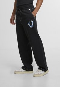 Pantalones de chándal negros con un corte relajado, que presentan costuras blancas, un diseño gráfico azul y bolsillos laterales. Llevados con zapatillas blancas.