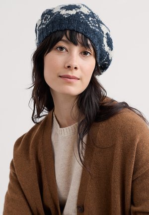 Pletena mornarsko plava i bijela beretka s teksturiranim uzorkom, uparena s smeđim kardiganom preko bež džempera, mekani i udobni materijali.