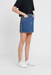 Kvinna som bär en blå denim minikjol med texten "Calvin Klein Jeans" på sidan, svart kortärmad t-shirt och vita sneakers, står med händerna i fickorna.