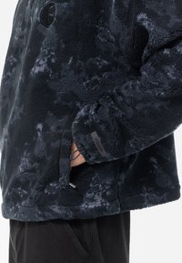 Fleecejakke i mørk marineblå med tie-dye mønster, featuring sidelommer, elastiske mansjetter, og et logo merke på brystet. Myk tekstur.