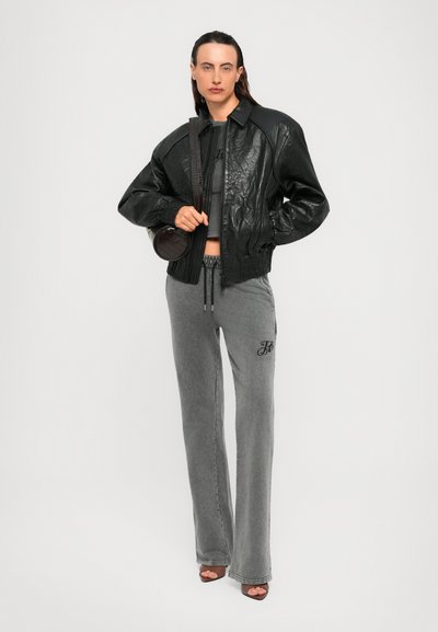 Persona che indossa una giacca di pelle nera oversize, pantaloni grigi a gamba larga con monogramma, tiene in mano una borsa rotonda nera e indossa tacchi aperti.