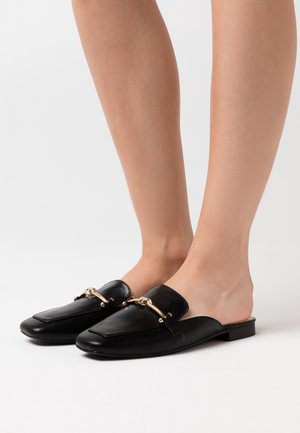 Mustat nahka loaferit, joissa on litteä pohja ja kultaketju koristeena edessä. Muotoilu on avoin kantapään kohdalla, mikä tekee niiden pukemisesta helppoa.