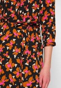 Närbild av en svart klänning med orange och rosa blommönster, ett knytbart tygbälte i midjan och en trekvartslång ärm med manschett.