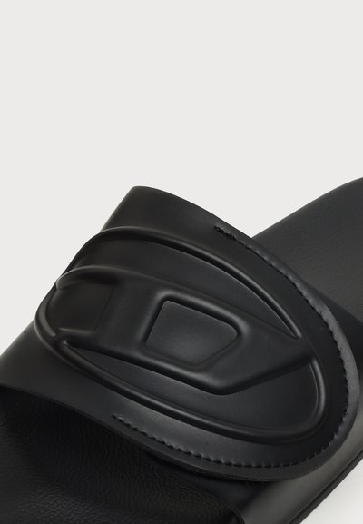 Sandale slide noire avec une large bride présentant un motif géométrique ovale en relief au centre.