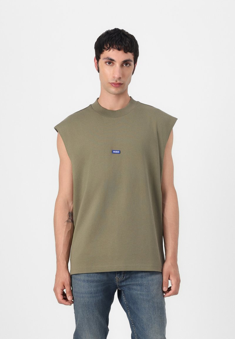 Camiseta sin mangas de color verde oliva, hecha de algodón, con un cuello redondo y una pequeña etiqueta de marca azul en el pecho. Textura suave.