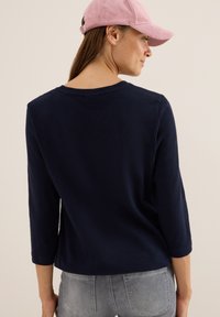 Kvinna som bär en marinblå långärmad skjorta, grå jeans och en rosa kordflorske baseballkeps, visad bakifrån och vänd något åt höger.