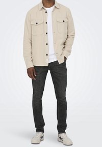 Camisa beige de botones con dos bolsillos en el pecho, combinada con jeans oscuros y zapatillas blancas. La camisa presenta una textura lisa y botones negros.