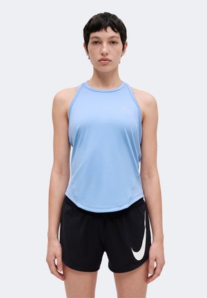 Jong persoon met kort haar draagt een lichtblauw mouwloos topje en zwarte sportshorts met een wit swoosh-logo tegen een effen lichte achtergrond.