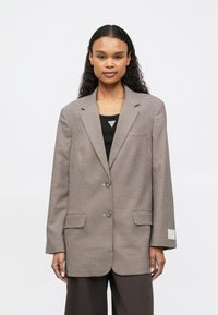 Femme avec de longs cheveux bouclés portant un blazer à carreaux marron sur un top noir et un pantalon large foncé, se tenant devant un fond blanc.